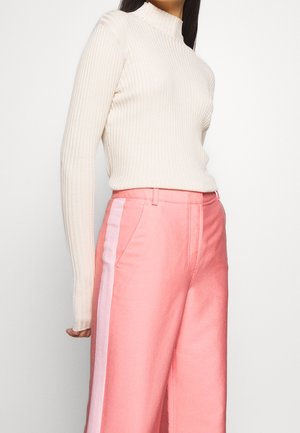 Femme portant un haut à manches longues côtelé crème et un pantalon taille haute rose avec une bande latérale rose clair, debout devant un fond blanc.