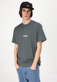 Tricou gri închis cu mâneci scurte și guler rotund, având textul logo alb pe piept. Purtați cu blugi din denim blue.