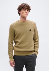 Νέος άνδρας με σγουρά μαλλιά, φορώντας μπεζ πλεκτό πουλόβερ με μικρό μαύρο κυκλικό λογότυπο και σκούρα γκρι παντελόνια, με τα χέρια στις τσέπες.