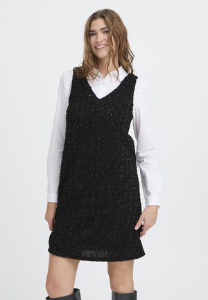 Jeune femme portant une robe sans manches noire texturée par-dessus une chemise blanche à manches longues, souriante sur un fond clair uni.