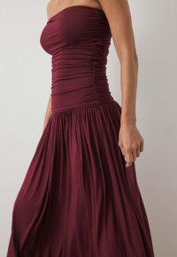 Burgundia rihmata maxi kleit, millel on volangiga rind, vöödiga lõige ja voolav seelik, mis on valmistatud pehmest, kergest kangast ja millel on peen tekstuur.