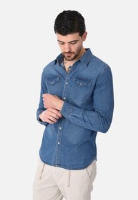 Camicia di jeans azzurro chiaro, maniche lunghe, con bottoni sul davanti, due tasche sul petto e orlo curvo. Texture morbida e design del colletto classico.