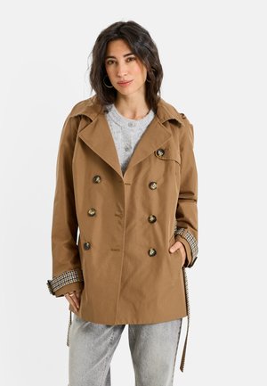 Femme portant un manteau marron à double boutonnage par-dessus un pull gris et un jean gris clair, debout avec une main dans sa poche sur fond blanc.