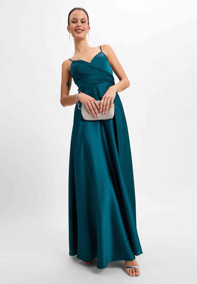 Tealfarbenes Satin-Maxikleid mit tailliertem Oberteil und fließendem Rock, mit dünnen Trägern und herzförmigem Ausschnitt. Hält eine silberne Clutch.