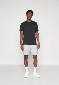 Schwarzes Baumwoll-T-Shirt mit Logo auf der Brust, kombiniert mit hellgrauen Sportshorts und weißen Sneakers. Das Modell steht vor einem schlichten Hintergrund.