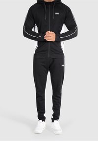 Svart zip-up hoodie med vita sidodetaljer, parat med matchande svarta joggers. Har en huva, elastiska ärmslut och vita sneakers.