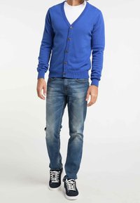 Blå bomullskardigan med V-hals, knappstängning fram och ribbade ärmslut, matchad med bleka blå jeans och svarta sneakers med vita snören.