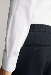 Camicia bianca a trama con polsini abbottonati accanto a pantaloni blu scuri con tasca posteriore e dettaglio con bottone.