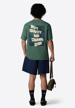Camisa de manga corta verde con un gran texto gráfico en la parte posterior que dice "DESAFÍA LA GRAVEDAD Y EL SENTIDO COMÚN." Combinada con pantalones cortos azul marino.