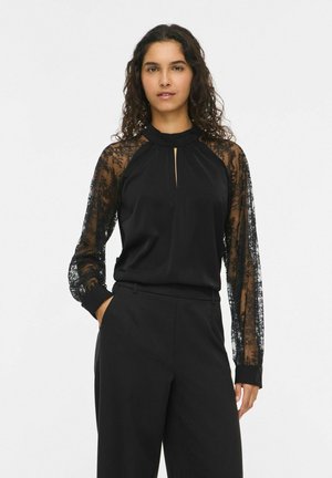 Une femme porte un chemisier noir aux manches en dentelle transparente, avec une petite fente à l’encolure, assorti à un pantalon noir sur un fond blanc uni.