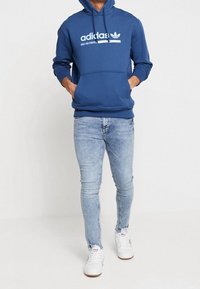 Hoodie en coton bleu avec une poche kangourou à l'avant, logo Adidas blanc imprimé sur la poitrine, associé à un jean skinny délavé et des baskets blanches.