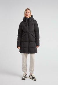 Kvinde står i en sort polstret frakke med hætte, hvide joggers og sort-hvide sneakers foran en ensfarvet baggrund.