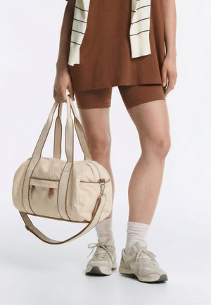 Persoon in bruine korte broek en top houdt een beige sporttas vast, draagt witte sokken en lichtgrijze sneakers tegen een effen achtergrond.