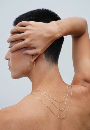 Goldschmuck umfasst gestapelte Ketten in unterschiedlichen Längen, ein schlanker Armreif und Creolen, die alle eine glatte, reflektierende Oberfläche aufweisen.