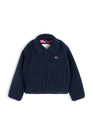 Niño con pelo largo y rubio lleva una chaqueta azul marino sobre un jersey rojo, vaqueros de pierna ancha color azul claro y zapatillas blancas, sonriendo y sosteniendo la chaqueta abierta.