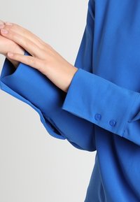 Händer justerar manschetten på en långärmad blå skjorta med två blå knappar på manschetten mot en ljusgrå bakgrund.