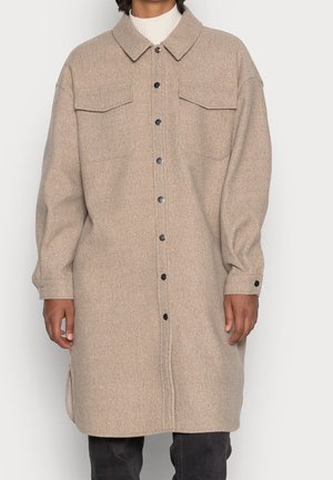 Beżowy długi płaszcz na guziki z klapkowymi kieszeniami na piersi i kołnierzem, noszony na białym golfie i ciemnych spodniach.