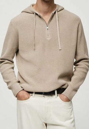 Jersey beige de punto con cremallera hasta la mitad y capucha, combinado con pantalones blancos y cinturón negro, manos metidas en los bolsillos contra un fondo liso.