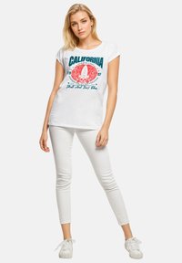 White cotton T-shirt with teal and coral graphic text and design featuring palm trees. Paired with white skinny pants and white sneakers.