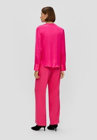 Helle rosa Satinbluse mit langen Ärmeln und Manschetten, kombiniert mit passenden rosa weiten Hosen und schwarzen High-Heels.