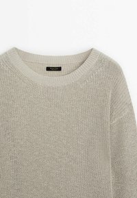 Pull en maille gris clair avec un col rond, présentant un tissage lâche et des poignets côtelés. Étiquette visible au niveau du col.