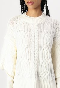 Maglione bianco lavorato a maglia con motivi a treccia testurizzati, colletto a girocollo a coste e maniche ampie. Il filato morbido e spesso aggiunge profondità e calore.