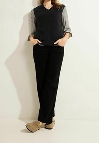 Maglione nero a collo a V sopra una camicia a maniche lunghe a righe, abbinato a pantaloni neri a gamba larga e zoccoli beige. Nessun motivo o dettaglio visibile.