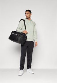 Sort duffelbag med mesh-detaljer, Champion-logo, båret af en person i en lysegrøn hættejakke og sorte bukser, der står foran en neutral baggrund.