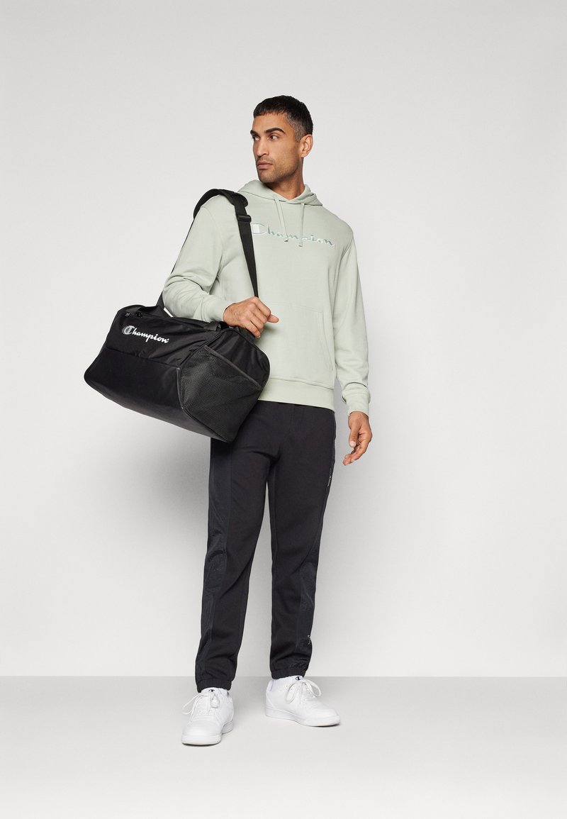 Sac à duffle noir avec des accents en maille, logo Champion, porté par une personne en sweat à capuche vert clair et pantalon noir, se tenant contre un fond neutre.