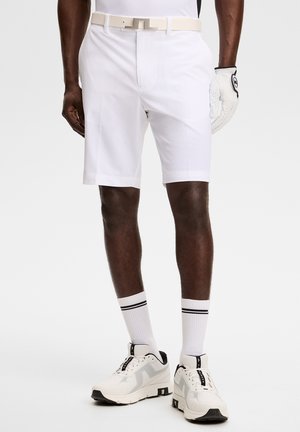 Mann trägt weiße Golfshorts, weißen Gürtel, weißen Golfhandschuh, weiße Socken mit schwarzen Streifen und weiße Sportschuhe, steht auf grauem Hintergrund.