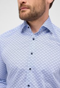 Hellblaues Button-Up-Hemd mit einem Muster aus kleinen dunklen Kreisen, mit einem Kentkragen und dunklen Knöpfen. Baumwollstoff mit glatter Textur.