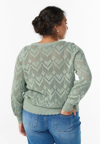 Gaiši zaļš adīts džemperis ar zigzaga rakstu, ribotiem piedurknēm un apakšmalu, brīvā piegriezumā ar atvērtu diegu dizainu. Apvienots ar zilām džinsām.