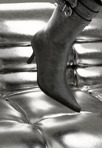 Black faux leather ankle boot with a pointed toe and a slim heel, featuring a studded strap around the ankle, resting on metallic surface.