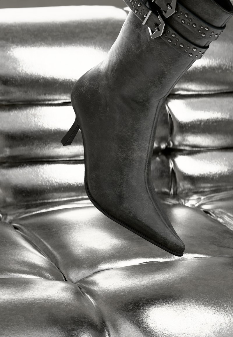 Black faux leather ankle boot with a pointed toe and a slim heel, featuring a studded strap around the ankle, resting on metallic surface.