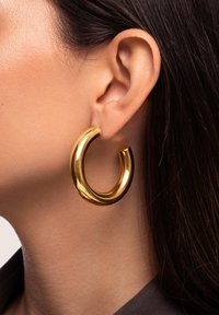 Orecchini a cerchio in oro con un design morbido e arrotondato, caratterizzati da una finitura lucida e una forma spessa e curvata, appesi all'orecchio di un modello.