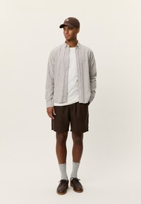 Ριγέ πουκάμισο με μακριά μανίκια και κουμπιά, λευκό μπλουζάκι, καφέ σορτς, γκρι κάλτσες, σκοτεινά παπούτσια και καφέ καπέλο, στέκεται μπροστά σε ουδέτερο φόντο.