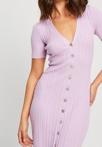 Femme portant une robe ajustée, côtelée, de couleur violet clair, avec des manches courtes et une rangée de boutons à l'avant allant de l'encolure à l'ourlet.