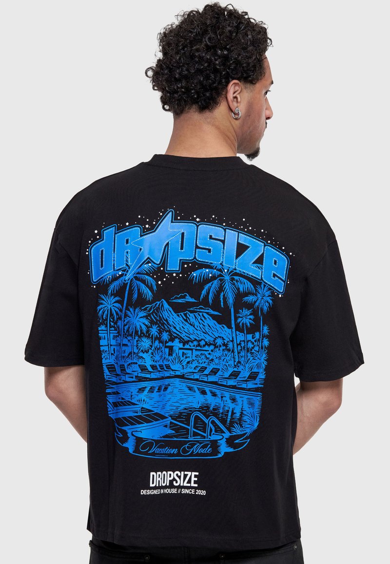 Homme aux cheveux bouclés portant un t-shirt noir avec un motif bleu représentant une scène au bord de la piscine, des palmiers, des montagnes et le texte « dropsize » dans le dos.