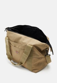 Khaki kanva kantokassi, jossa on kaksoiskangaskahvat, vetoketjusuljin ja musta vuorattu sisustus. Sisältää etutaskun, jossa on logopainatus.