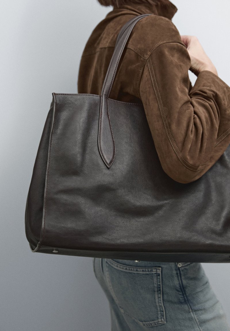 Geräumige dunkelbraune Leder-Tote-Tasche mit glatter Textur, spitz zulaufenden Griffen und minimalistischem Design, die am Arm getragen wird.