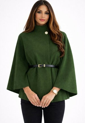 Femme portant un manteau cape vert foncé ceinturé avec des manches larges, un pantalon noir et une montre-bracelet dorée, debout devant un fond blanc.