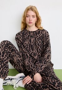 Brūns un melns leopardu raksta mājas apģērba komplekts; garām piedurknēm tops un brīvi bikses no mīksta, teksturēta auduma. Baltas sporta kurpes ar melniem akcentiem.