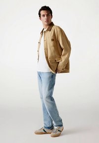 Beżowa kurtka dżinsowa z guzikiem z przodu i kieszenią na piersi, biała koszulka, jasnoniebieskie dżinsy oraz beżowe trampki z zielonymi akcentami.