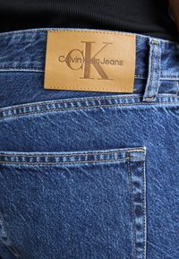 Traperice s kožnom oznakom u boji pijeska na struku koja sadrži utisnute natpise "Calvin Klein Jeans" i "CK." Plava tkanina s vidljivim detaljima šivanja.