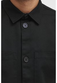 Black, woven shirt featuring a collar, two chest pockets, and black buttons. The fabric has a smooth texture with a subtle sheen.