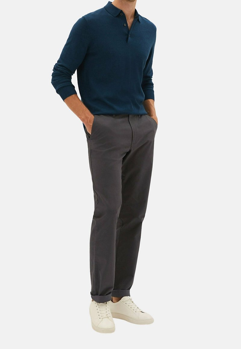 Homme portant un polo bleu marine à manches longues, un chino gris foncé avec revers et des baskets blanches à lacets, debout les mains dans les poches.