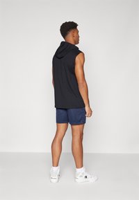 Schwarze ärmellose Kapuzenjacke mit Kapuze, kombiniert mit navyblauen Shorts. Modell trägt weiße Sportschuhe mit dezenten Akzenten und strukturiertem Material.