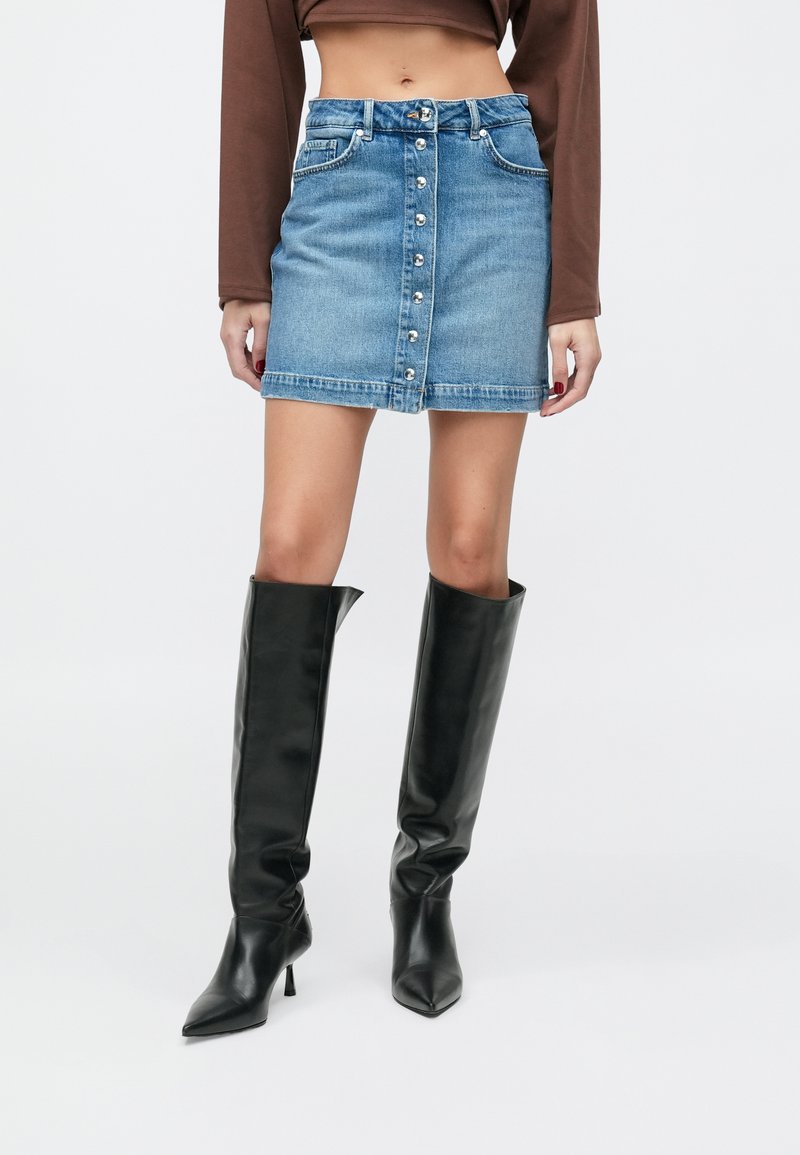 Falda vaquera en azul claro, con cierre frontal de botones, dos bolsillos y diseño ajustado. Combinada con botas negras altas y holgadas.