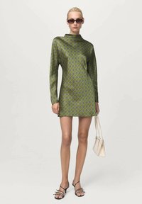 Vestido verde con estampado, de mangas largas, cuello alto y un diseño geométrico. Combinado con sandalias negras con tiras y un pequeño bolso blanco.