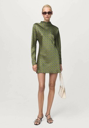 Abito verde a motivo con maniche lunghe, collo alto e un design geometrico. Abbinato a sandali neri con cinturini e una piccola borsa bianca.